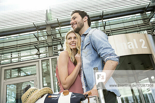 Lächelnde Frau  die ihren Freund umarmt  während sie mit einem Gepäckwagen aus dem Flughafenterminal ankommt