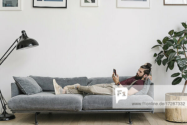 Mann benutzt ein Mobiltelefon  während er auf dem Sofa im Wohnzimmer liegt