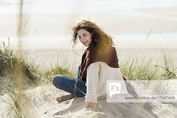 Lächelnde Frau schaut über die Schulter  während sie mit gekreuzten Beinen am Strand sitzt