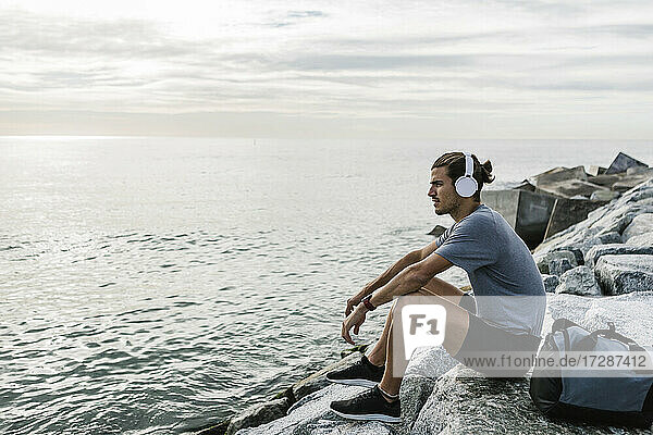 Sportler mit Kopfhörern  der auf einem Felsen am Meer sitzt und die Aussicht betrachtet