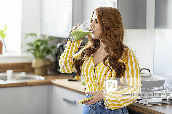 Rothaarige Frau trinkt gesunden Milchshake  während sie in der Küche zu Hause steht