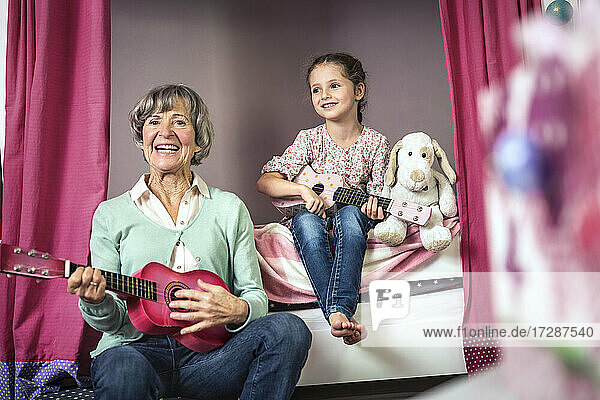 Lächelnde Großmutter und Enkelin mit Gitarre im Schlafzimmer sitzend
