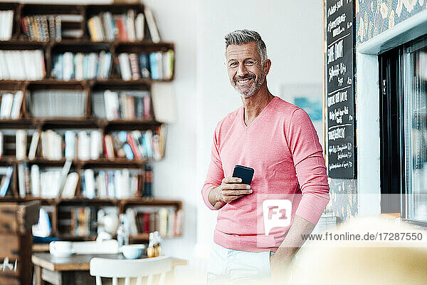 Reifer männlicher Unternehmer  der ein Smartphone hält  während er mit den Händen in den Taschen in einem Café steht