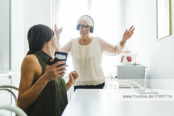Mittlere erwachsene Frau sitzt mit Smartphone und schaut ihrer Großmutter beim Tanzen zu  während sie zu Hause über Kopfhörer Musik hört
