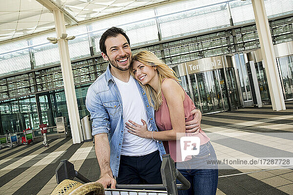 Glückliches junges Paar mit Gepäcktransportwagen im Flughafenterminal