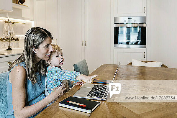 Junge zeigt auf Laptop  während er auf dem Schoß der Mutter sitzt und in der Küche arbeitet