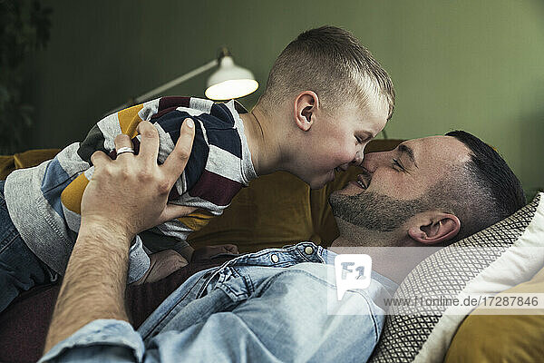 Glücklicher Vater  der seinen Sohn umarmt  während er zu Hause auf dem Sofa im Wohnzimmer liegt