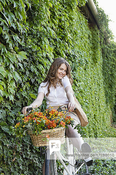 Lächelnde junge Frau auf dem Fahrrad sitzend vor einer grünen Efeuhecke