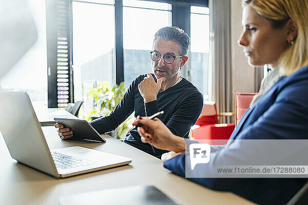 Älterer Geschäftsmann  der auf seinen Laptop schaut  während er mit einer Mitarbeiterin in einem kreativen Büro diskutiert