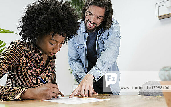 Lächelnder Geschäftsmann  der eine Kollegin bei der Unterzeichnung eines Dokuments an einem Schreibtisch im Büro begleitet