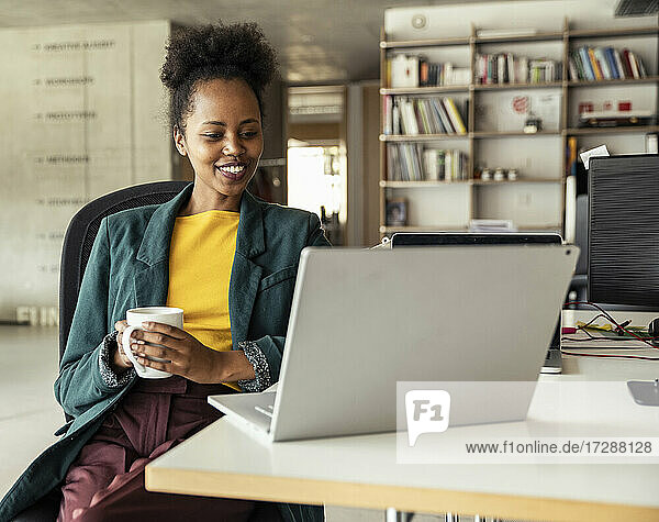 Lächelnde weibliche Fachkraft  die während einer Videokonferenz über einen Laptop im Büro Kaffee trinkt