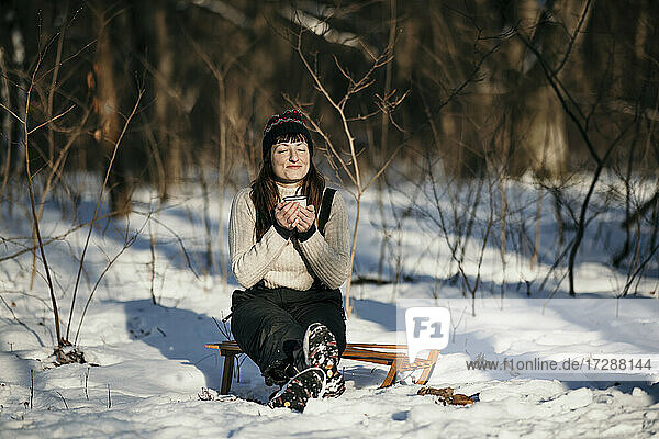 Frau hält Becher und sitzt auf einem Schlitten im Wald