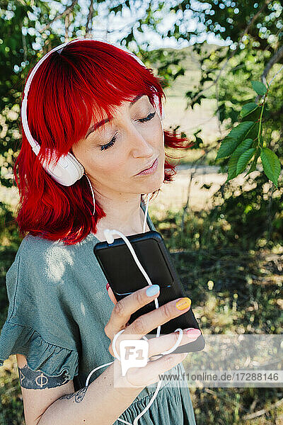 Beautiful woman with eyes closed holding mobile phone while listening music in front of tree