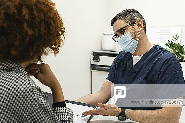 Männlicher Zahnarzt mit Gesichtsschutzmaske diskutiert mit einer Patientin in der Klinik über ein Zahnschema