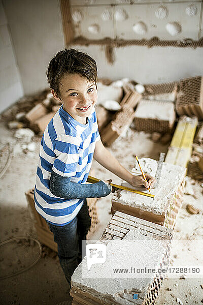 Lächelnder Junge  der während einer Renovierung an einem Haus steht und Ziegel misst