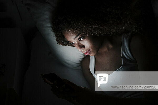 Frau  die ihr Smartphone benutzt  während sie auf dem Bett in einem dunklen Raum zu Hause liegt