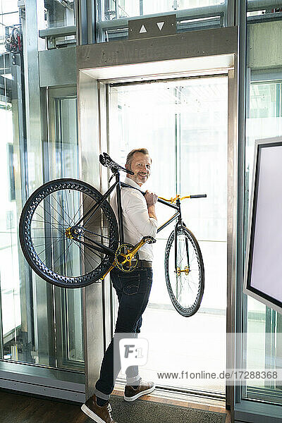 Lächelnder Geschäftsmann mit Fahrrad auf der Schulter beim Verlassen des Büros