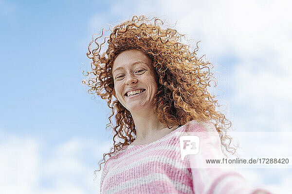 Fröhliche rothaarige Frau mit lockigem Haar steht unter dem Himmel
