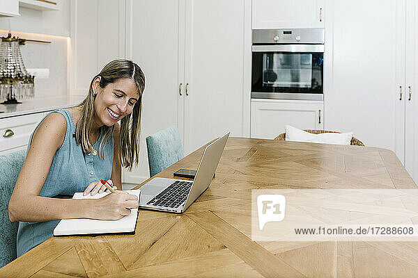 Lächelnde Freiberuflerin  die am Esstisch in der häuslichen Küche in ihr Tagebuch schreibt