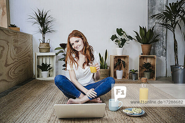 Lächelnde junge Frau sitzt im Schneidersitz und trinkt Saft vor einem Laptop auf dem Boden zu Hause