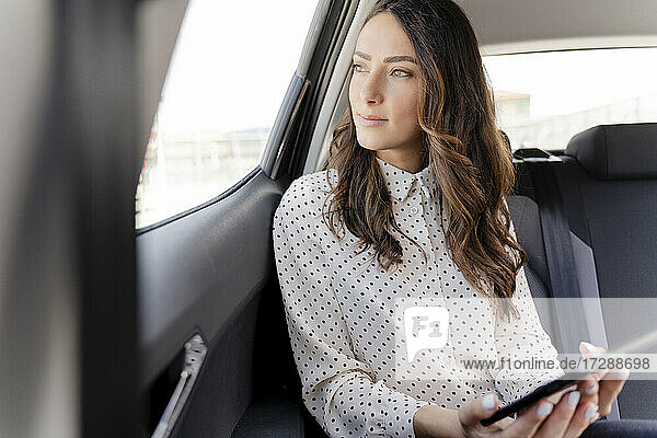Geschäftsfrau mit digitalem Tablet  die aus dem Fenster schaut  während sie im Auto sitzt