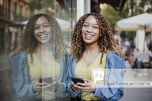 Lächelnde Frau mit Smartphone  die sich an eine Glaswand lehnt