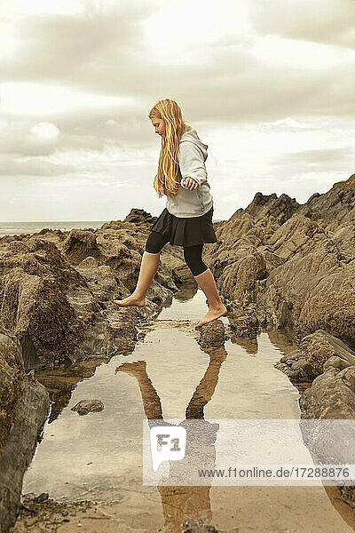 Blondes Mädchen hüpft auf einer Felsformation über einem Gezeitenbecken am Strand