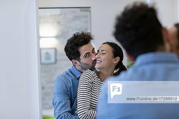 Boyfriend kissing girlfriend on cheek in front of mirror at home