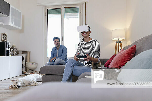 Junge Frau spielt mit virtueller Realität  während Mann und Haustiere zu Hause sitzen