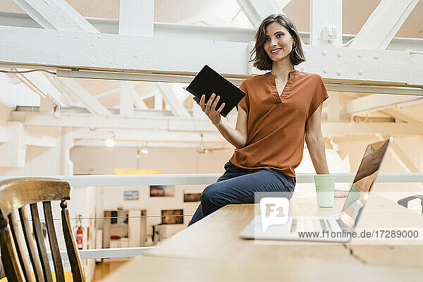 Smiling beautiful woman with diary sitting on desk in creative office looking away