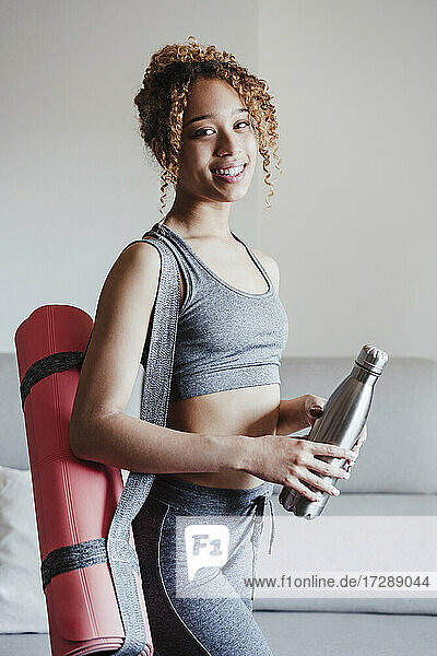 Young woman with exercise mat and bottle standing by sofa in living room
