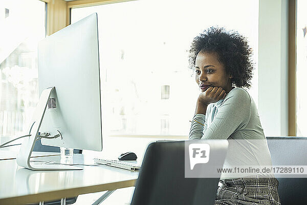 Weibliche Fachkraft mit Hand am Kinn  die einen Computer auf dem Schreibtisch im Büro benutzt
