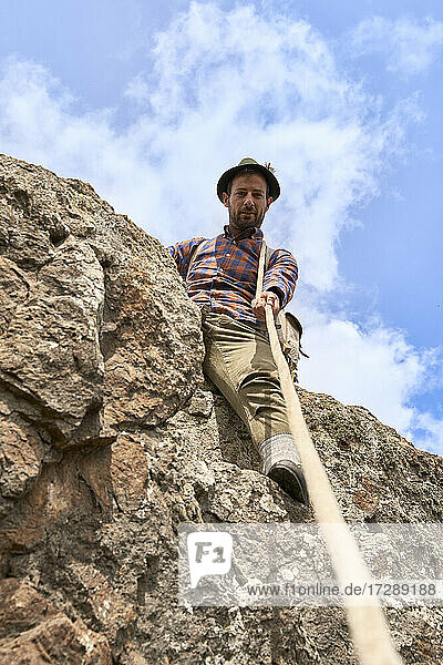 Male mountaineer holding rope while sitting on rock during sunny day
