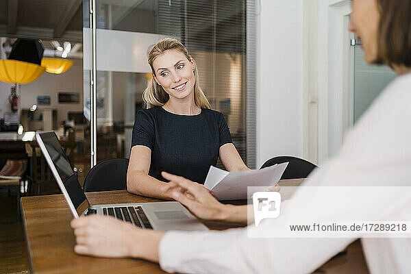 Lächelnde Geschäftsfrau  die auf einen Laptop schaut  während sie mit einem Kollegen an einem Schreibtisch im Büro sitzt und diskutiert