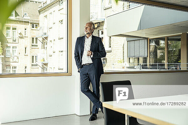 Male professional holding coffee cup while leaning near glass window at office