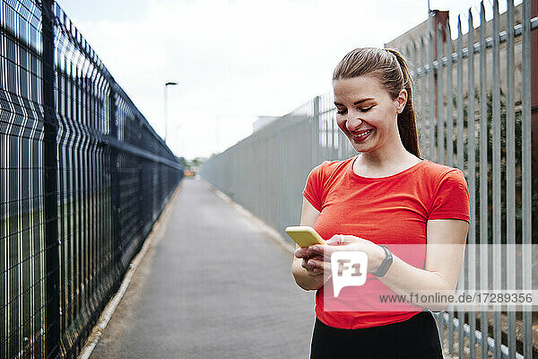 Smiling fit woman text messaging on smart phone on footpath