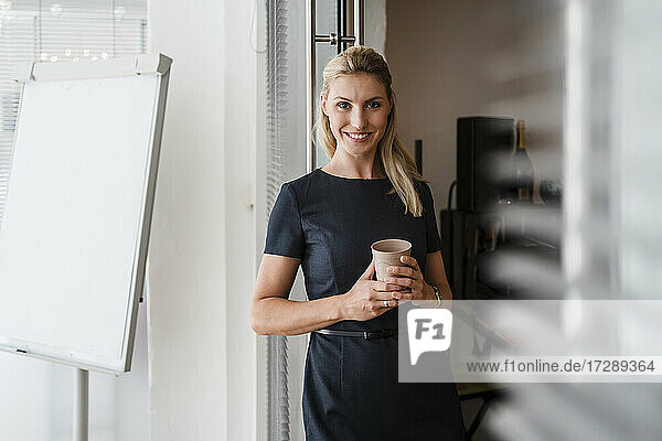 Lächelnde Unternehmerin mit wiederverwendbarem Kaffeebecher an der Tür im Büro stehend