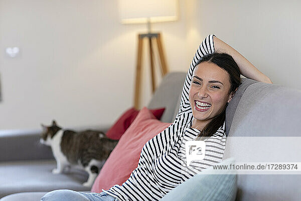 Fröhliche junge Frau mit getigerter Katze im Hintergrund auf dem Sofa zu Hause
