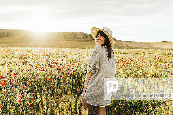 Smiling woman wearing hat and shirt standing at poppy field during sunny day