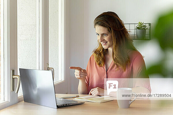 Lächelnde Geschäftsfrau  die während eines Videogesprächs auf einen Laptop im Büro zu Hause zeigt