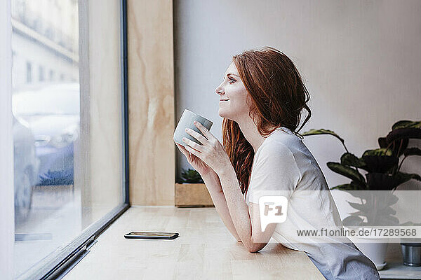 Smiling woman having coffee while leaning on window sill at home