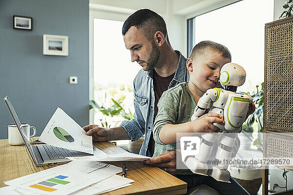 Junge spielt mit einem sitzenden Roboter  während ein Geschäftsmann eine Grafik zu Hause analysiert