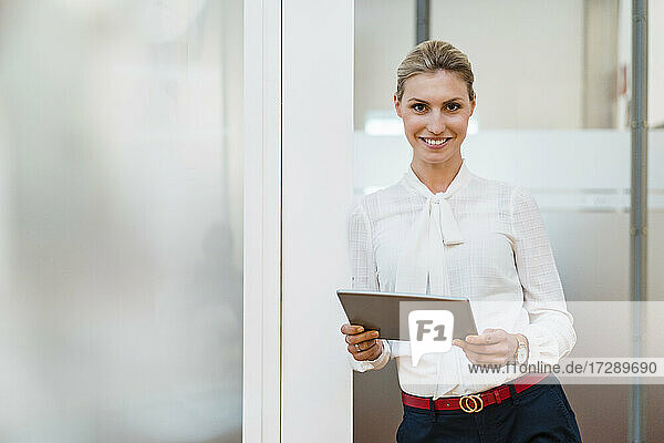 Lächelnde weibliche Fachkraft mit digitalem Tablet  die an der Tür im Büro lehnt