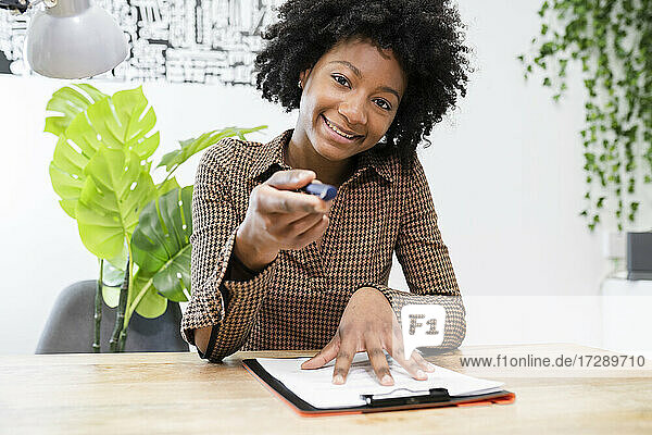 Lächelnde Geschäftsfrau  die einen Stift hält und mit einem Klemmbrett am Schreibtisch im Büro sitzt