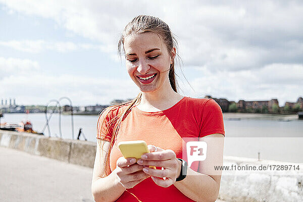 Smiling fit woman text messaging on smart phone in front of sky