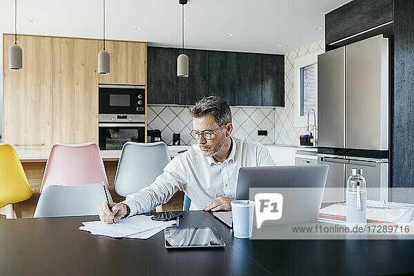 Male freelance worker wearing eyeglasses writing on documents at table in home office