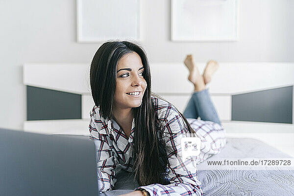 Lächelnde Frau  die wegschaut  während sie mit einem Laptop im Schlafzimmer liegt