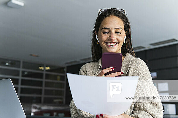 Lächelnde junge Geschäftsfrau mit Dokument  die eine Voicemail auf einem Smartphone im Büro sendet