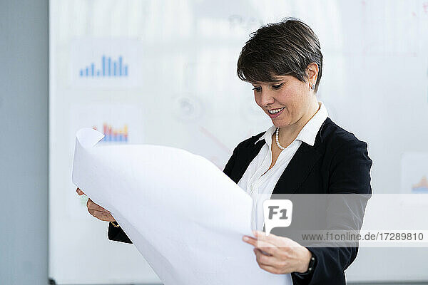 Lächelnde weibliche Fachkraft  die eine Papiertabelle bei der Arbeit im Büro analysiert