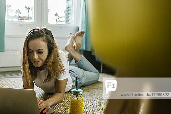 Lächelnde Frau  die einen Laptop benutzt  während sie zu Hause auf dem Teppich liegt